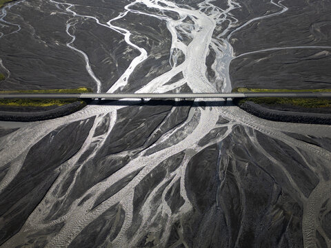 Aerial view of a bridge crossing a complex braided river system with dark volcanic sediment and glacial water channels in Iceland.