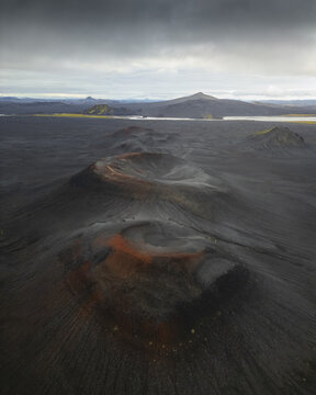 Aerial view of volcanic craters with black ash slopes and reddish soil under a dramatic overcast sky in Iceland.