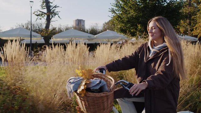 Smiling young woman rides bicycle past dry plants in autumn park