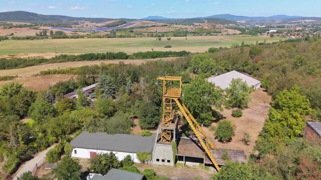 Drone view of the historic Tiribesi-Akna coal mine headframe amid woodland and farmland near B&aacute;tonyterenye, Hungary, with the M&aacute;tra hills and village beyond.