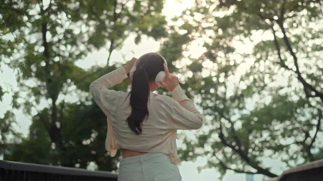 Happy young Asian woman dancing joyfully outdoors while listening to music with wireless headphones, raising arms and moving freely in green park, rear view of carefree female enjoying rhythm, freedom