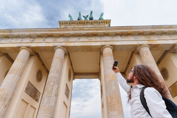 Young man with long hair and a backpack captures a photograph of the majestic Brandenburg Gate using his mobile device while exploring the historic center of Berlin Germany. © Katie Chizhevskaya