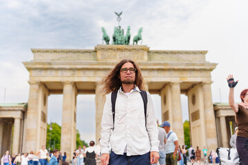 Long-haired man wearing glasses and a light-colored button-down shirt stands in front of the iconic Brandenburg Gate located in the heart of Berlin, Germany during the day. © Katie Chizhevskaya