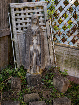 Weathered stone statue of the Virgin Mary praying in a garden leaning against a rustic wooden trellis lattice fence