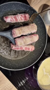 ​Close-up video of meat rolls wrapped in bacon sizzling and frying in a hot skillet pan with spatula and fork, preparing home cooked dinner.
