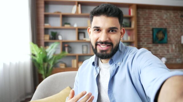 A happy smiling man speaks during an online video call on a smartphone in a POV shot at home. He looks into the camera, chats cheerfully with friends, and relaxes on a cozy sofa in a living room.