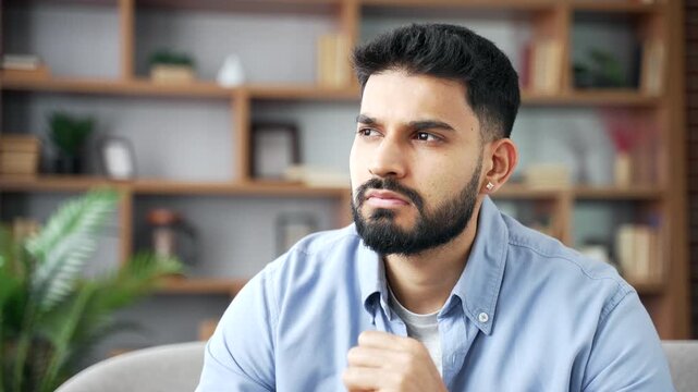 Close-up of a serious bearded man looking thoughtfully aside while sitting indoors on comfortable couch. A focused male stays calm, thinking deeply and reflecting on his ideas in modern home apartment
