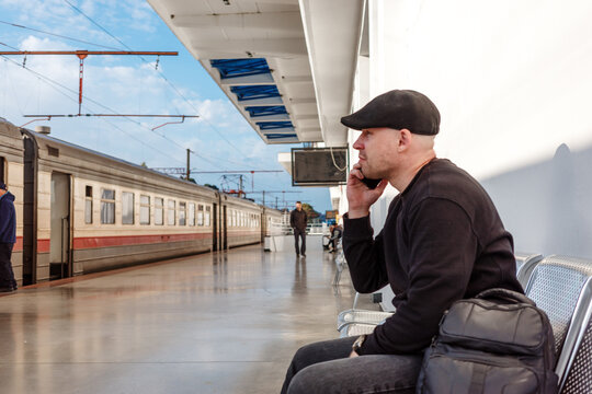 Man talking on phone while waiting for train at railway station platform, passenger sitting with backpack beside passenger train during rail travel and public transport journey.