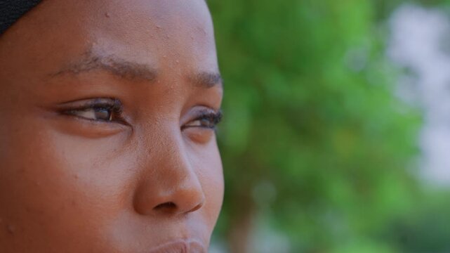 Close up view shows side profile of young adult female subject. Her skin texture details are visible with natural lighting and woman wears earring. background is softly blurred greenery
