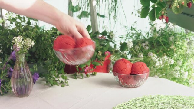 Woman places bowls of homemade strawberry sorbet on a table in a lush balcony garden and adds spoons for serving. Fresh summer dessert and outdoor dining lifestyle scene