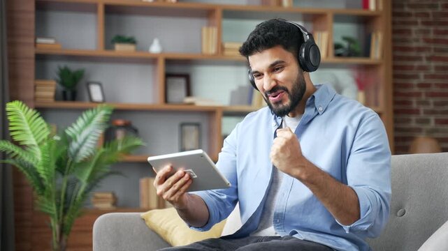 Excited male wearing wireless headphones watching live sports broadcast on digital tablet, sitting on sofa in living room at home. Happy fan emotionally cheering, celebrating victory in a cozy room.