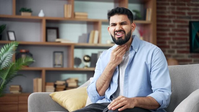 Male sitting on sofa at home, suffering from severe sore throat and holding neck with hands. Unhappy person experiencing painful throat inflammation, cold, flu, infection, and feeling unwell in room.