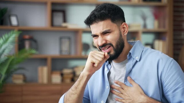 Close up of coughing man holding hand to chest while sitting on sofa at home. Unhappy male suffering from severe cold, flu, bronchitis, respiratory infection, stress, and feeling unwell in room.