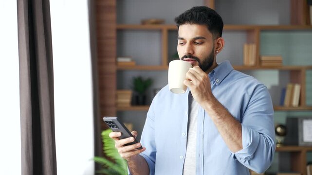 A relaxed bearded man standing by a window, holding a coffee cup and using a smartphone with a smile. A happy male drinks warm beverage and scrolls phone screen, enjoying a cozy morning at home.