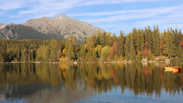 Scenic Mountain Lake with Autumn Forest Reflections in the High Tatras, Slovakia. The calm water reflects golden trees, rugged mountains, and a blue sky, creating a peaceful natural landscape 