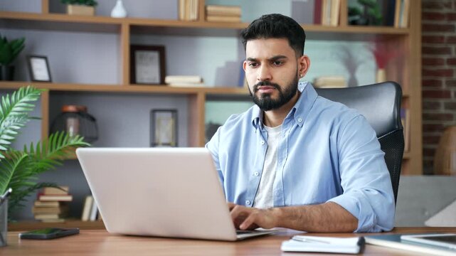 A thoughtful focused businessman uses a laptop and types on a keyboard at home. A pensive freelancer thinks deeply while working online, sits at a desk, and spends time in a modern home office.