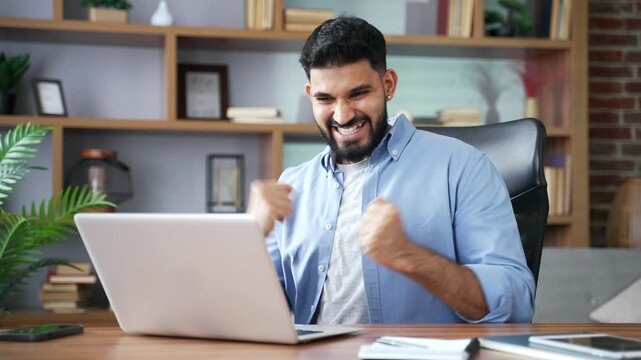 An excited happy man reads great news on a computer screen indoors. A thrilled businessman celebrates good results, reacts enthusiastically to a message, and sits at a desk in a home office.