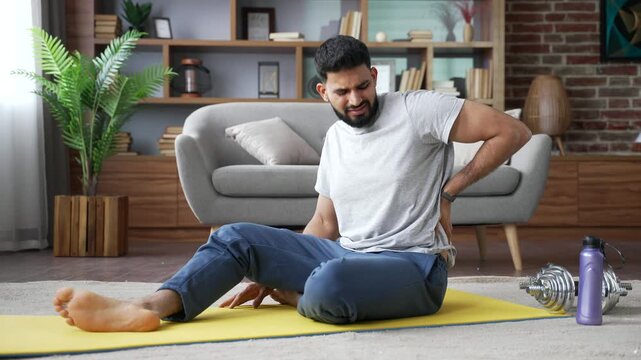 A man sitting on a mat on the floor at home suffers from back pain and muscle spasm after workout. He holds his painful flank with a hand to relieve lower spine discomfort and physical sports injury.