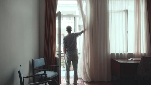 Man opening curtains to let bright morning sunlight flood room through balcony doorway, pulling sheer fabric aside for fresh start to day and natural light at home.