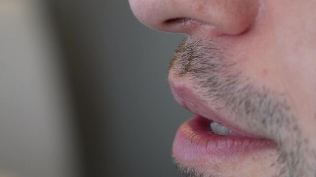 Side profile macro of a man&rsquo;s lips moving as he speaks, his nose and stubble in sharp focus against a soft grey background. A close up for talking, speech, pronunciation, lip reading, voice recording