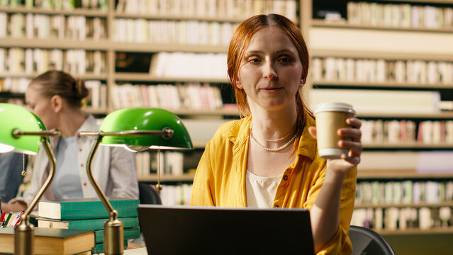Coffee, laptop and student as red-haired woman raises a takeaway cup while pausing at her computer. Study break, refreshment and campus routine connect coursework with a brief rest in a library.