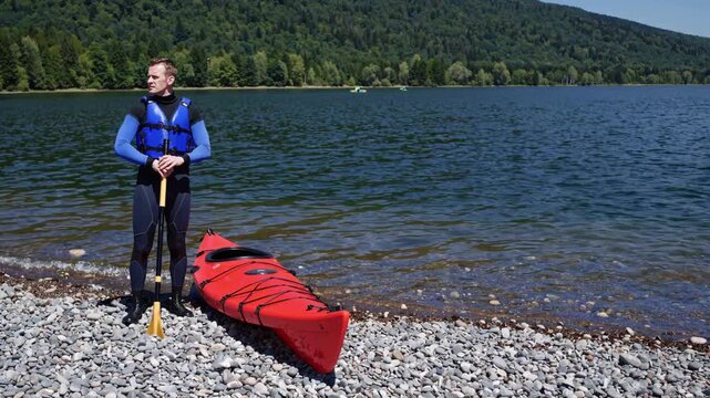 Man wearing life vest holds paddle upright next to red kayak on stony shoreline. Adventurous paddler looks out over quiet blue lake, enjoying peaceful nature during summer vacation journey.
