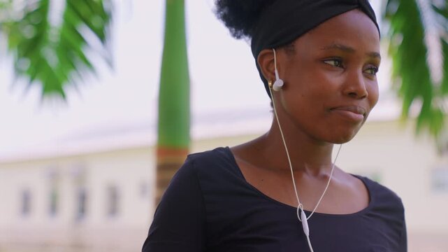 Young adult female listens to audio through white earbuds while standing outside. Woman wears black top with green foliage and out of focus building visible behind her