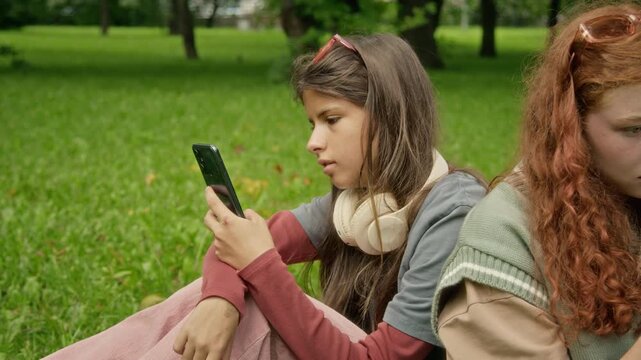 Caucasian teenage girl sitting on green grass outdoors, browsing smartphone near female friend. Suitable for youth mobile app marketing, social media connectivity, or digital lifestyle campaigns