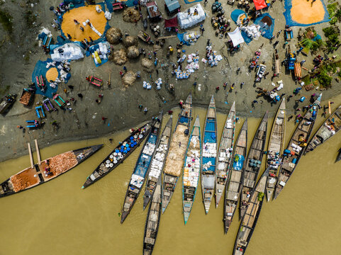 Kazipur, Rajshahi Division, Bangladesh - 24 August 2024: Aerial view of a riverbank market with wooden boats docked along the shore, workers loading sacks, and produce spread on the ground.