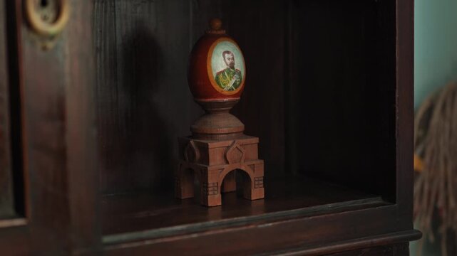 A wooden egg-shaped ornament featuring an image of Tsar Nicholas II. It sits on a wooden shelf in an old house.