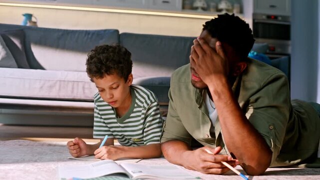 Exhausted father holding head helping son homework floor. Tired parenting home.