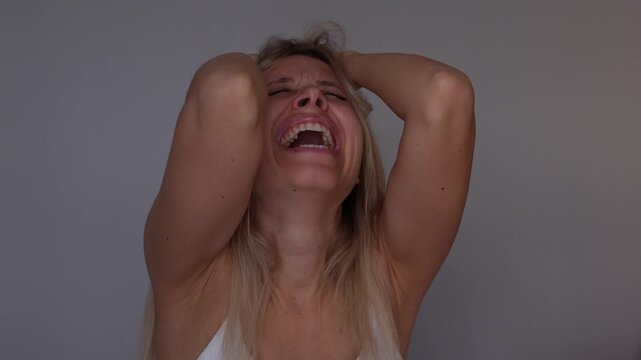 Young blonde woman holds her head with both hands and cries in overwhelming emotional pain, portraying intense psychological distress, helplessness and an inability to cope with unbearable feelings