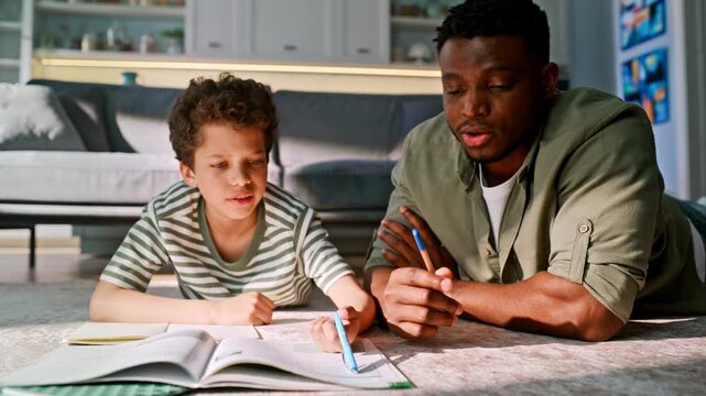 Father helping son homework lying living room floor. Dad teaching boy writing.