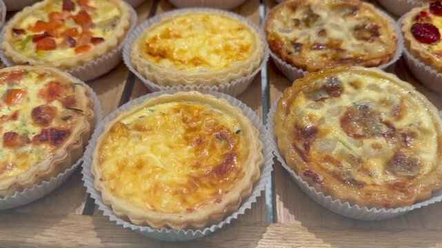 Small savory pies and French quiches with various fillings are arranged on a wooden showcase inside a bakery shop. Gourmet pastry display, bakery food retail, delicious catering assortment.