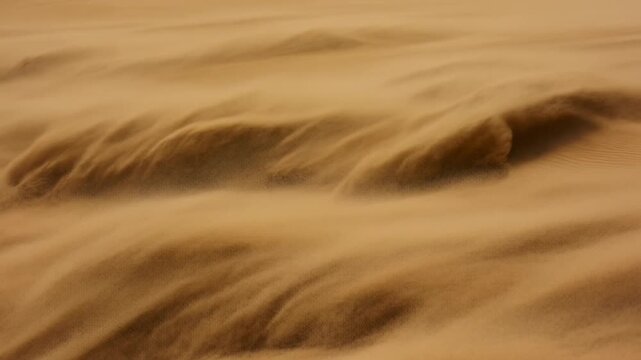 Windblown sand sweeps across golden desert dunes, reshaping rippled ridges