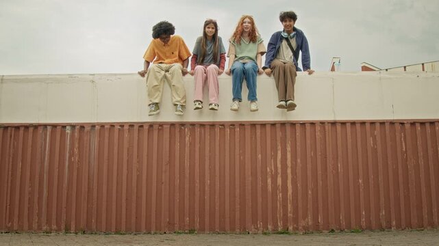 Portrait of multiethnic teenagers sitting on outdoor wall ledge, talking and dangling legs. Perfect for youth lifestyle, friendship and high school education campaigns