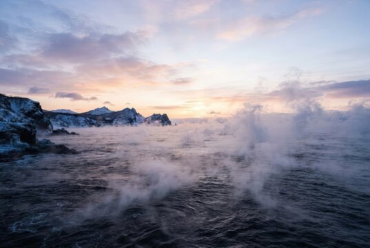 Sea smoke rising above cold ocean waters near snow-covered rocky shorelines under a morning sky.