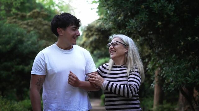 A Caucasian mother with glasses and her young adult son in a white T-shirt walk arm in arm towards the camera along a park path. They talk and smile as they pass trees and shrubs.