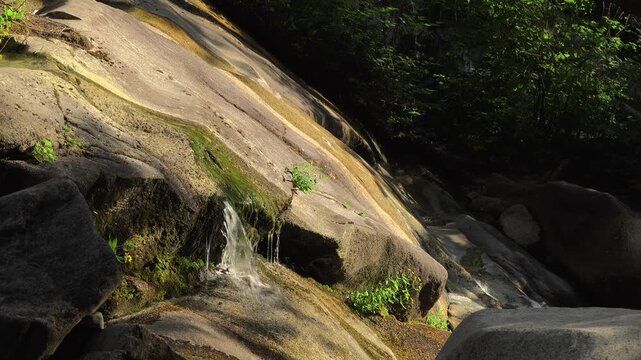 Close-up view of fresh water trickling and cascading over smooth, sunlit granite rock slabs at the bottom of Shannon Falls in Squamish, British Columbia. Small green plants grow on the stone.