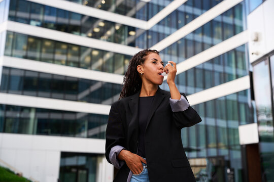 Asthma inhaler use outside a modern office tower. African businesswoman takes a dose of respiratory medication during her workday in a glass skyscraper courtyard.