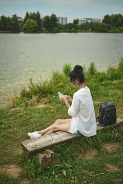 Woman sits on wooden bench by lake in evening. She wears white shirt and shorts. Black backpack is beside her. Trees and buildings can be seen in the background