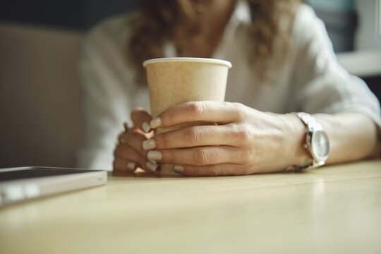A person enjoys a peaceful moment in a cozy cafe, holding a paper cup of coffee. Hands rest comfortably on a wooden table, showcasing a casual and relaxed atmosphere during midday