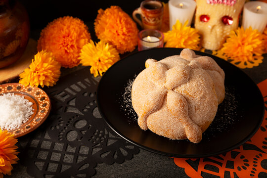 Traditional Mexican Pan de Muerto sweet bread on a black plate, placed on a Day of the Dead altar setup with cempasuchil marigold flowers and sea salt background.