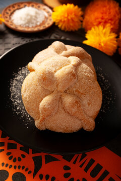 Vertical close-up of traditional Mexican Pan de Muerto sweet bread on a black plate, placed on a Day of the Dead altar setup with cempasuchil marigold flowers and sea salt background.