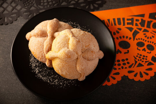 High angle shot of two traditional Mexican Pan de Muerto sweet breads on a matte black plate next to a festive orange papel picado paper cutout for Day of the Dead.