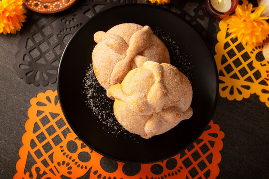 Mexican Pan de Muerto sweet breads on a matte black plate next to a festive orange papel picado paper cutout for Day of the Dead Ofrenda.