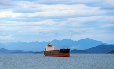 Large commercial cargo ship anchored at sea near a coast with misty blue mountains under a cloudy sky, representing maritime trade, global logistics, and shipping transportation © John_Doo78