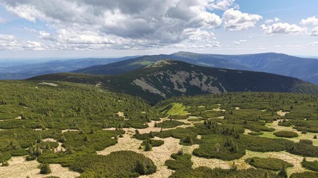 Breathtaking wide mountain panorama with sunlit alpine slopes, sparse shrubland, dark forested midground, and blue distant ridges beneath scattered clouds, calm outdoor atmosphere.