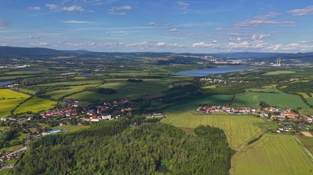 Lush rural panorama shows patchwork farmland, curved waterways, and scattered settlements across rolling hills, lit by bright summer sunlight and clear sky.
