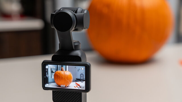 Camera stabilizer capturing a close-up of a large orange pumpkin on a table, with a blurred kitchen background and Halloween decorations visible